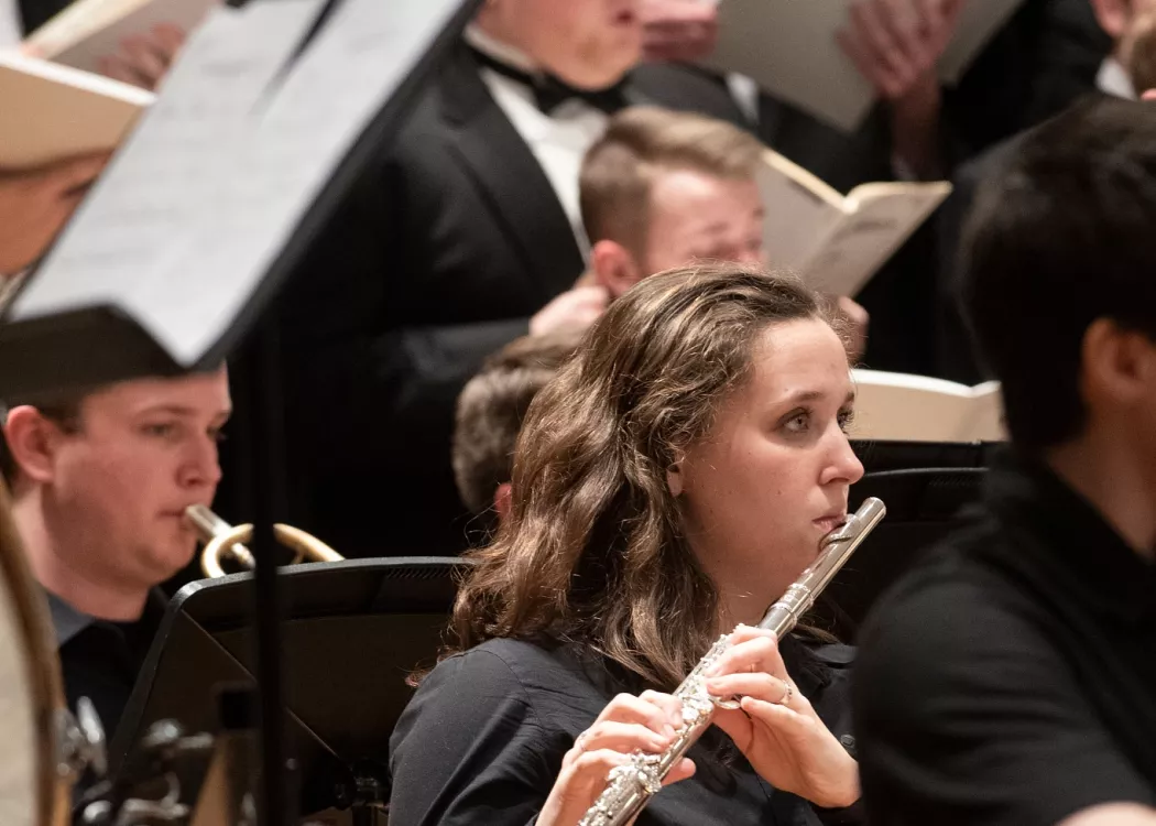 Student playing the flute with singers in the background at a music symphony concert.